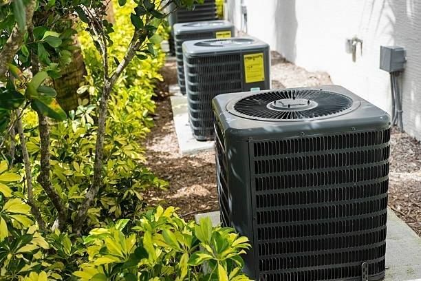 Air conditioning units lined up outdoors next to a building and greenery.