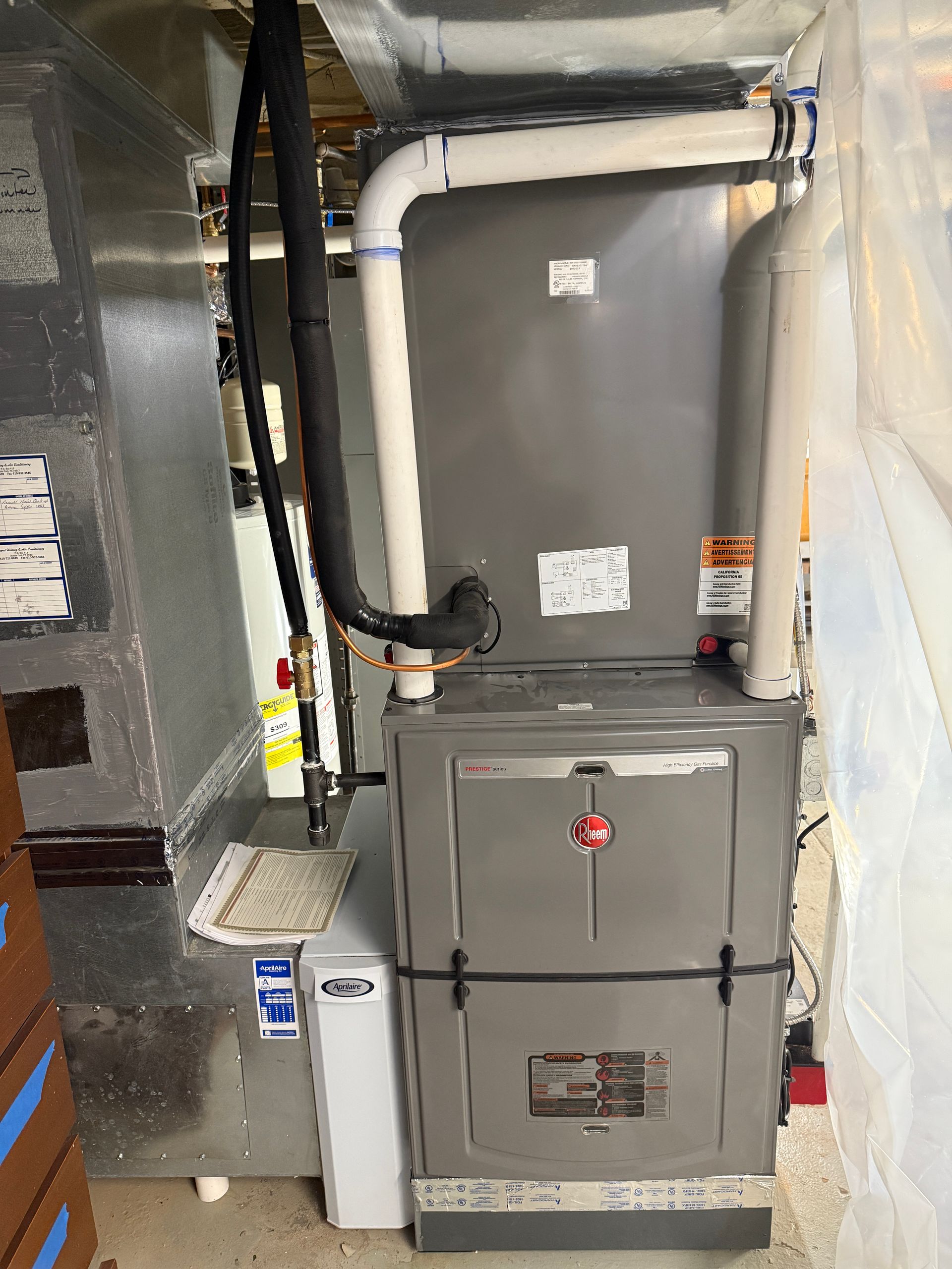 Furnace unit in a basement setting with ductwork and piping. Gray metal, white plastic, black insulated tubing.