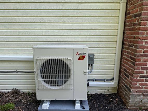 Mitsubishi heat pump unit outside a building with white siding and a brick wall.