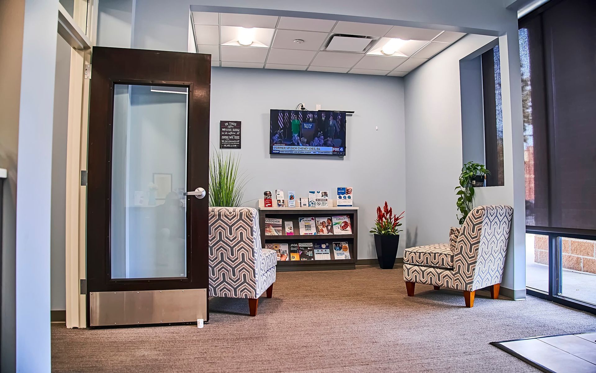 Waiting room with patterned chairs, wall TV, and glass entrance door in a bright office lobby
