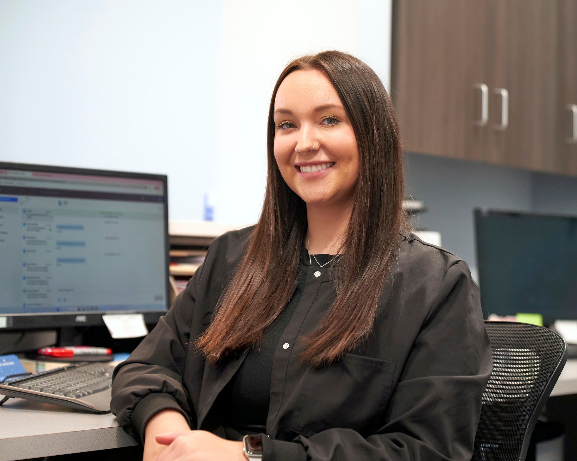 Woman smiling at a desk in an office, with a computer monitor and cabinets behind her.