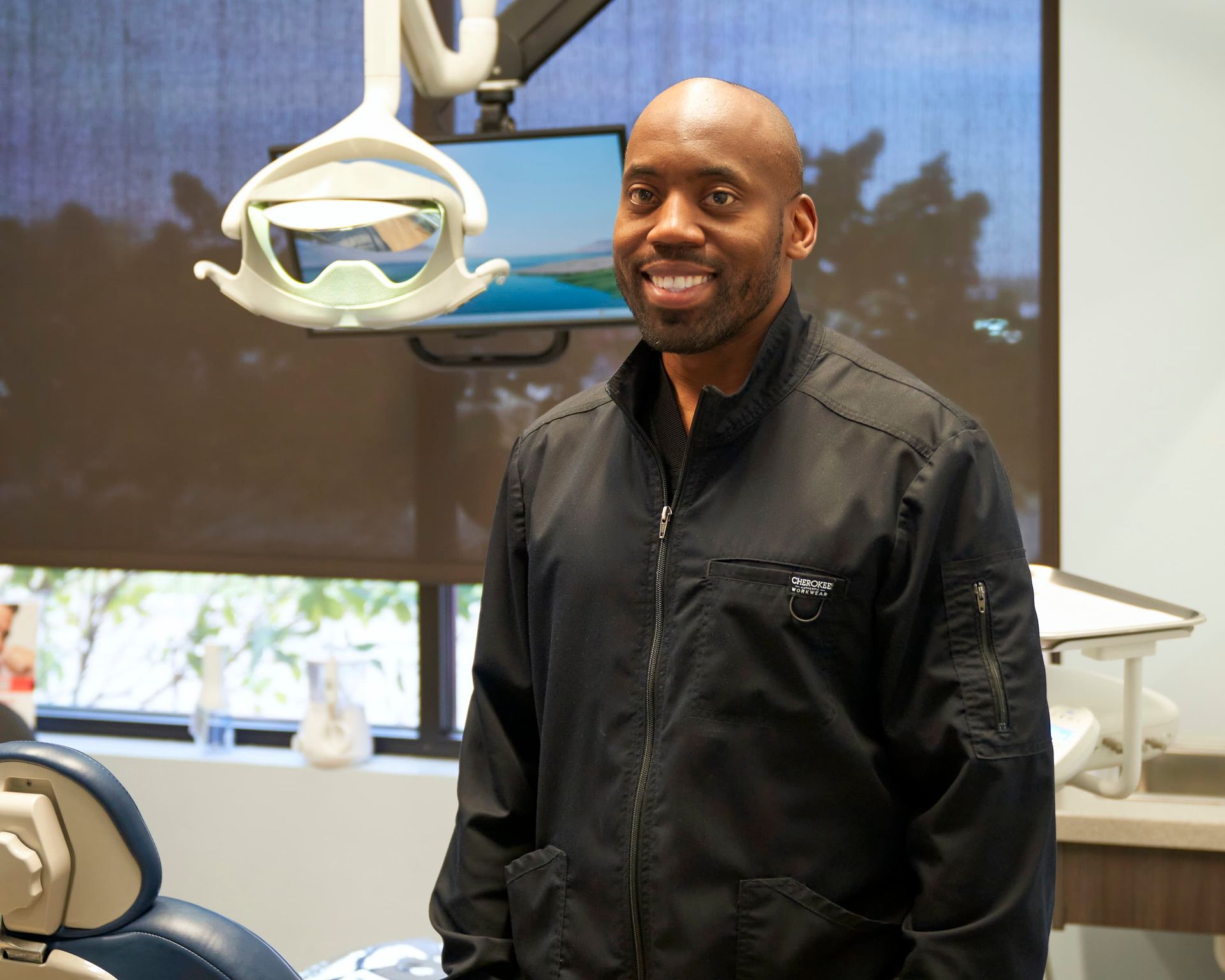 Man standing in a dental clinic beside a dental chair, smiling near a ceiling-mounted exam light