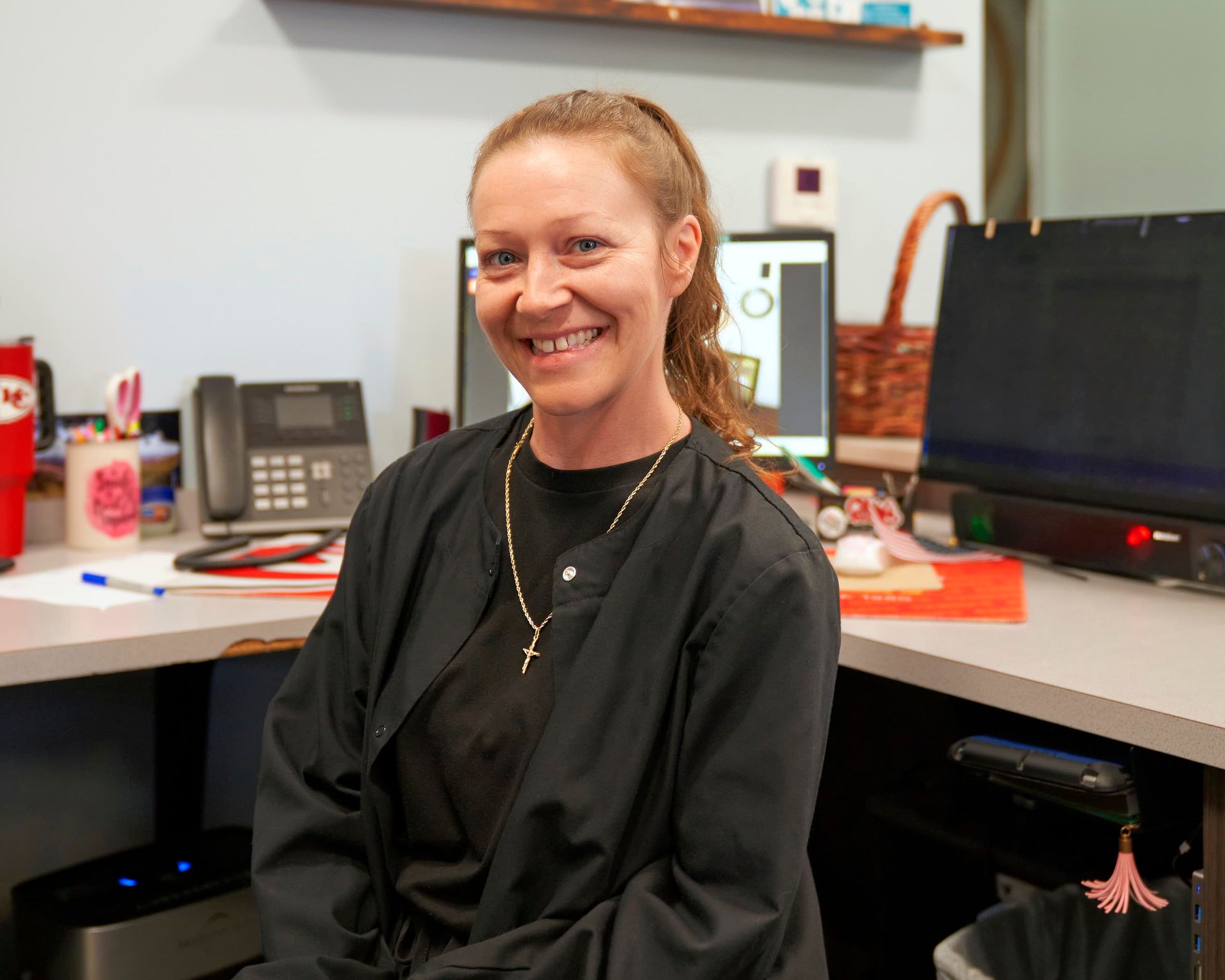 Smiling person in a black shirt seated at a desk in an office with computer monitors and office supplies.