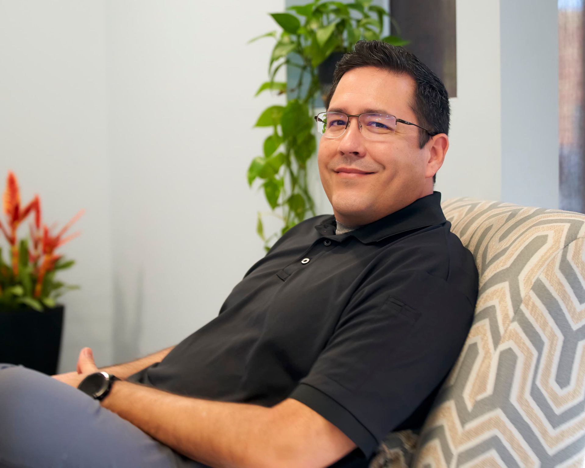 Man sitting on a patterned chair in an office, wearing a black shirt and smiling