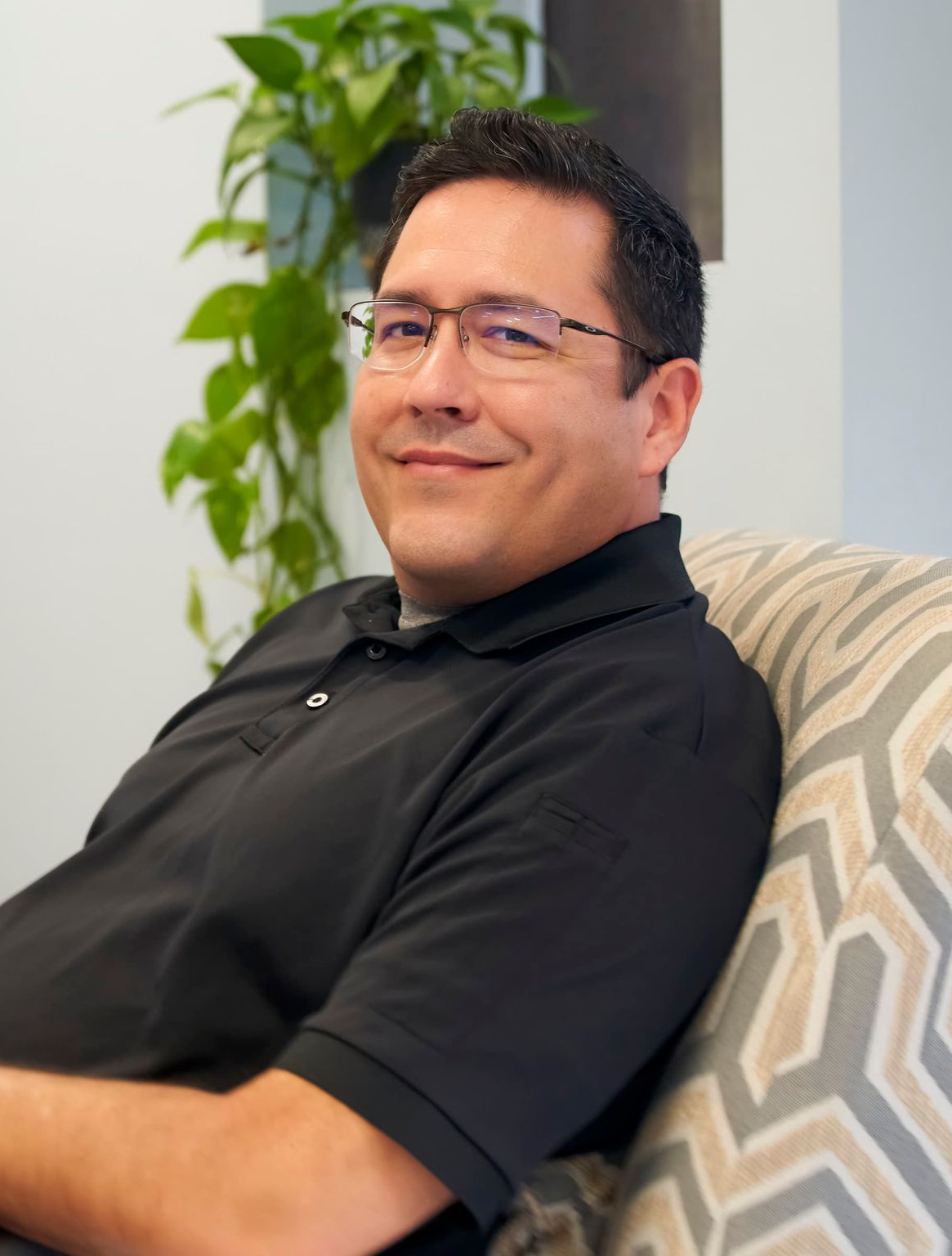 Man sitting on a patterned chair in an office, wearing a black polo shirt and smiling slightly.