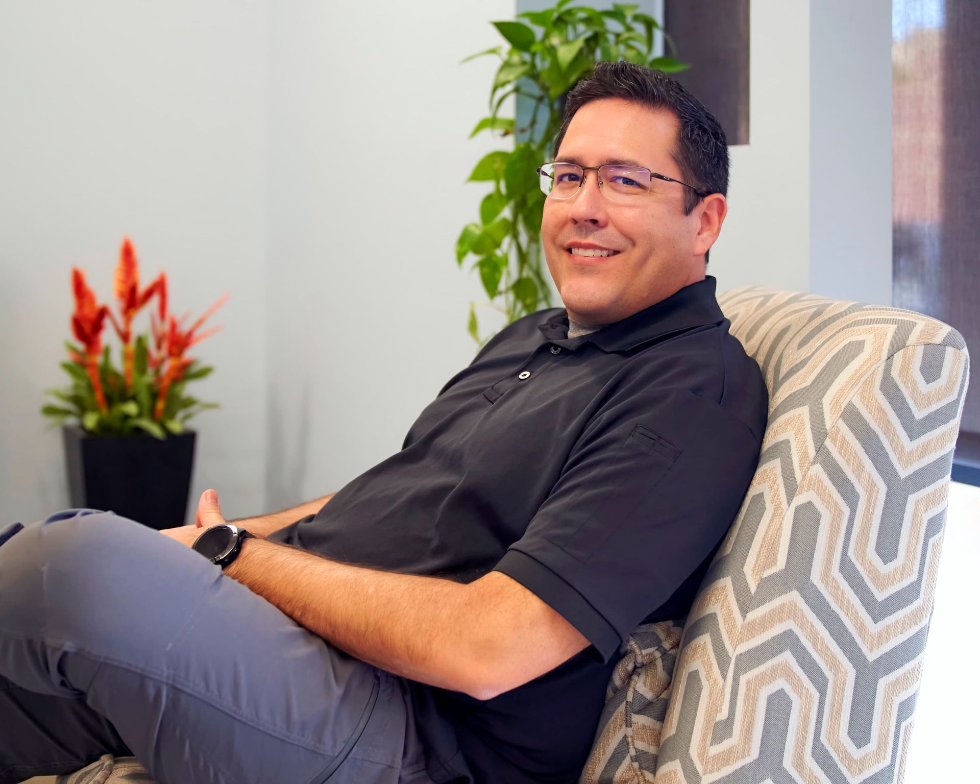 Man sitting in a patterned chair in an office, smiling and holding a device, with plants in the background.