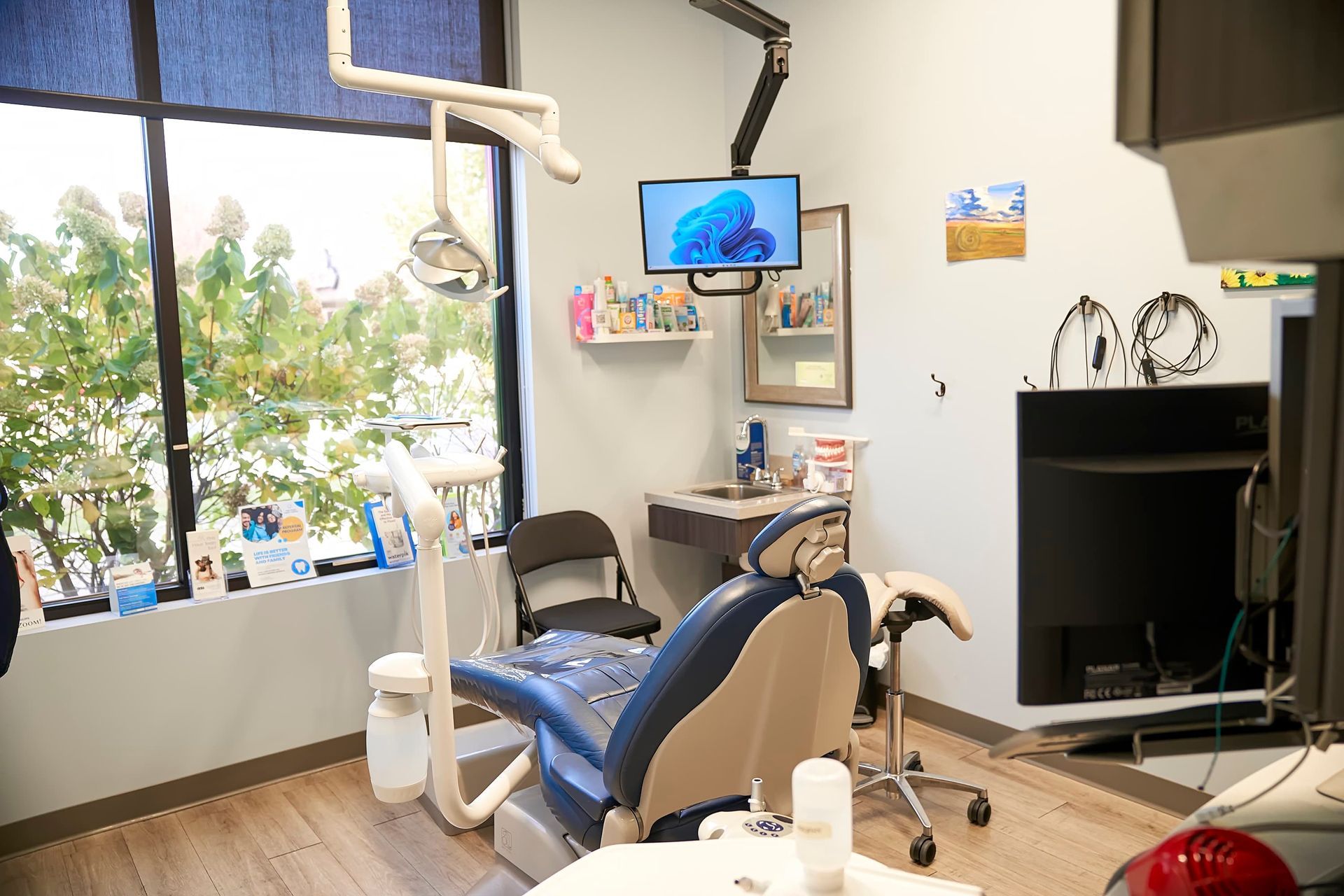 Dental office exam room with green chair, equipment, and large windows with outdoor view.