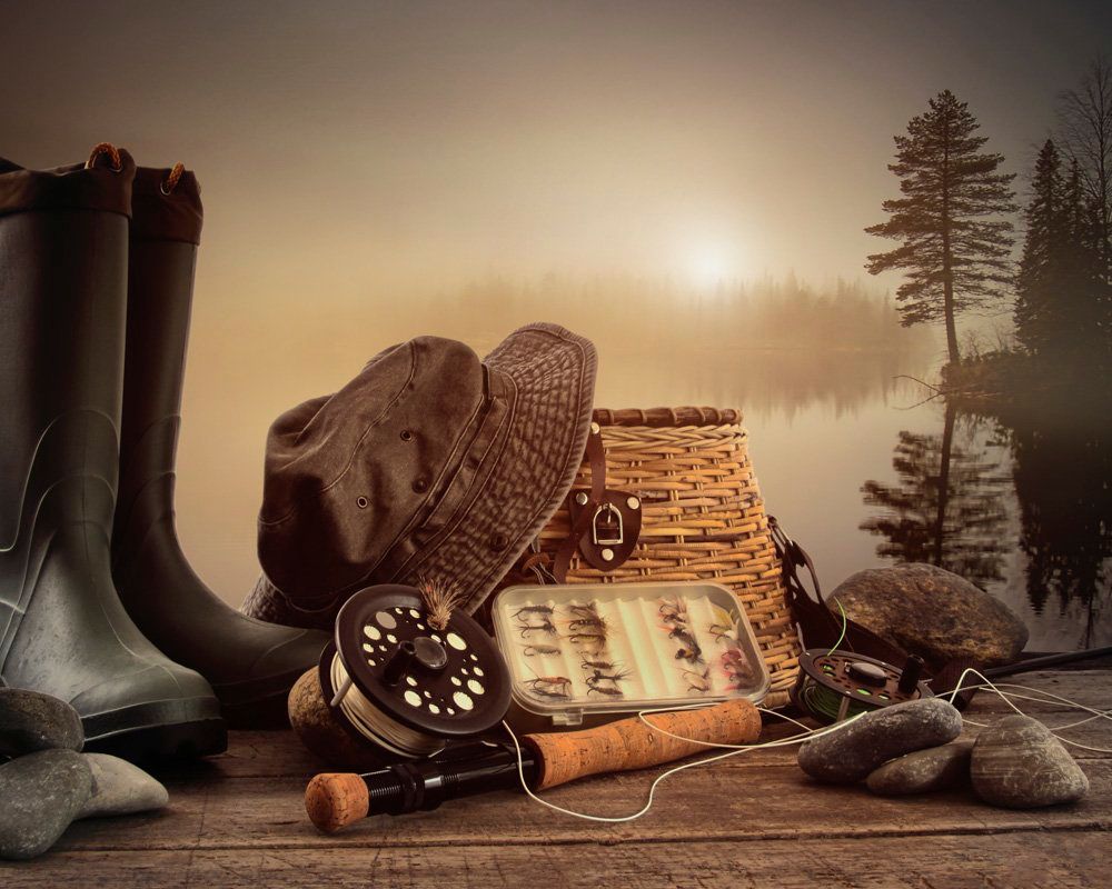 Fishing gear: boots, hat, creel, reel, fly box, and gloves on wooden surface with misty lake backdrop.