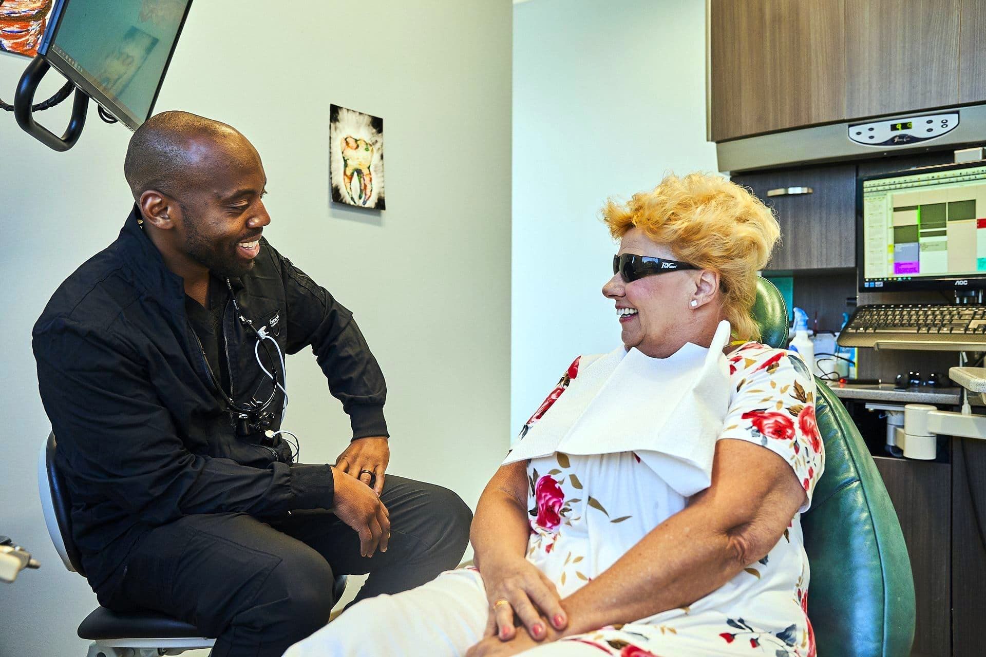 A dental professional converses with a patient in a dental office. Both are smiling.