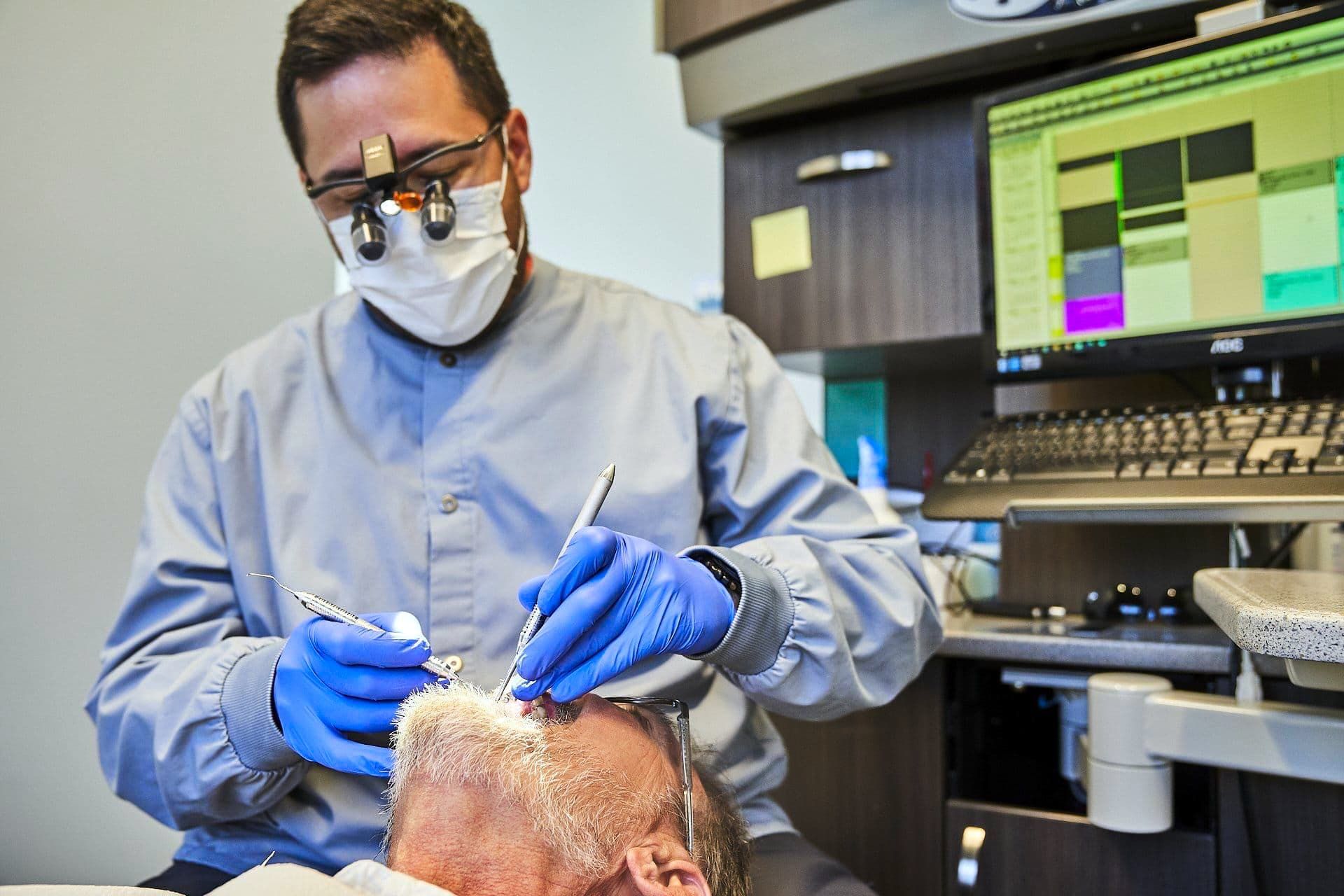 Dentist in scrubs, mask, and glasses working on a patient's head in a dental office.