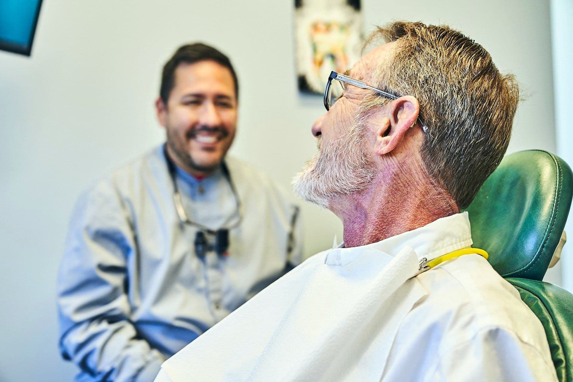 Dentist smiling at a patient in a dental chair. The patient wears a bib.