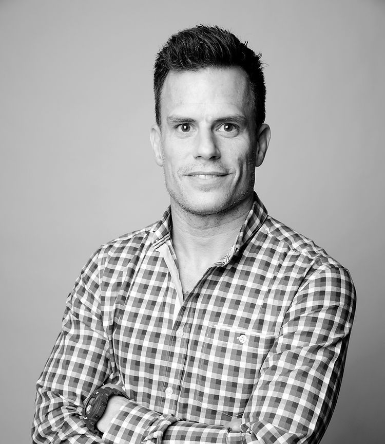 A person wearing a checkered button-down shirt with arms crossed, smiling slightly against a plain gray background.