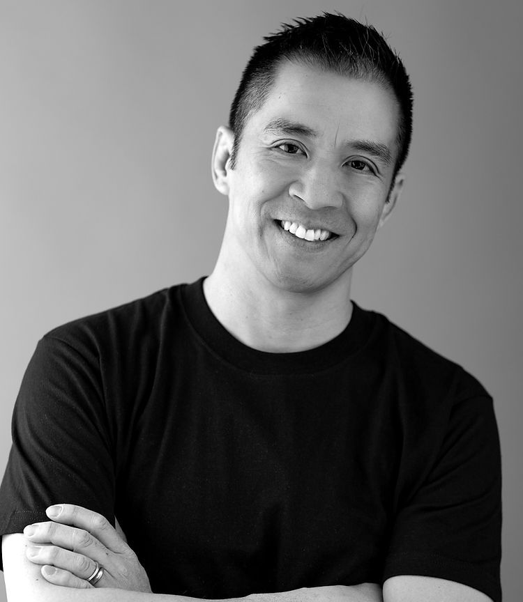 A smiling individual in a black t-shirt standing with arms crossed against a plain grey background.