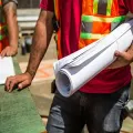 A worker in a red shirt and high-visibility vest holds a rolled-up set of blueprints at a construction site.