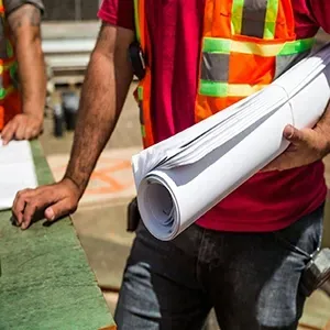 A worker in a red shirt and high-visibility vest holds a rolled-up set of blueprints at a construction site.
