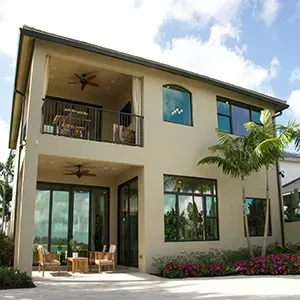 A two-story beige house with a covered patio on the first floor and a matching balcony above, featuring dark-framed windows.