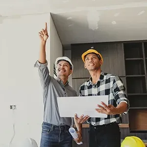 Two construction workers, one pointing to a ceiling and the other holding a laptop in an unfinished room.