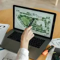 A person uses a pen to point at a landscape design plan on a laptop screen, with markers on a wooden desk.