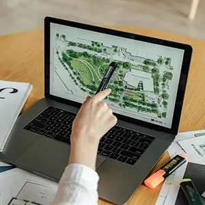 A person uses a pen to point at a landscape design plan on a laptop screen, with markers on a wooden desk.