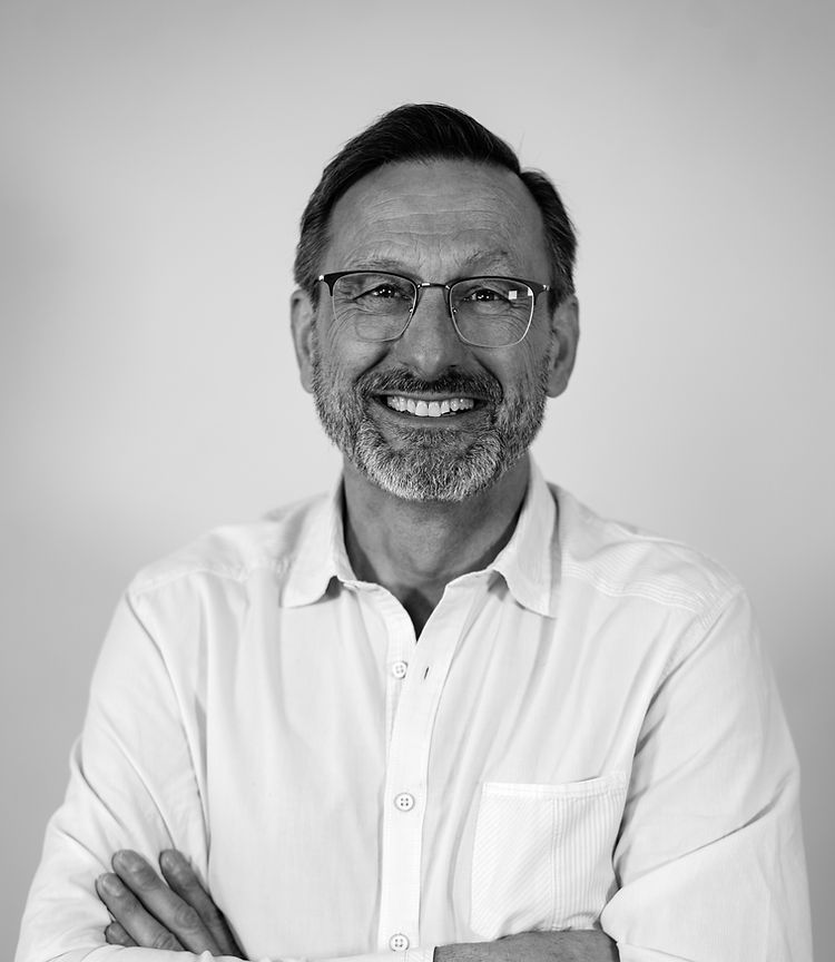 A smiling person with glasses and a beard wearing a button-down shirt, photographed from the chest up against a plain wall.
