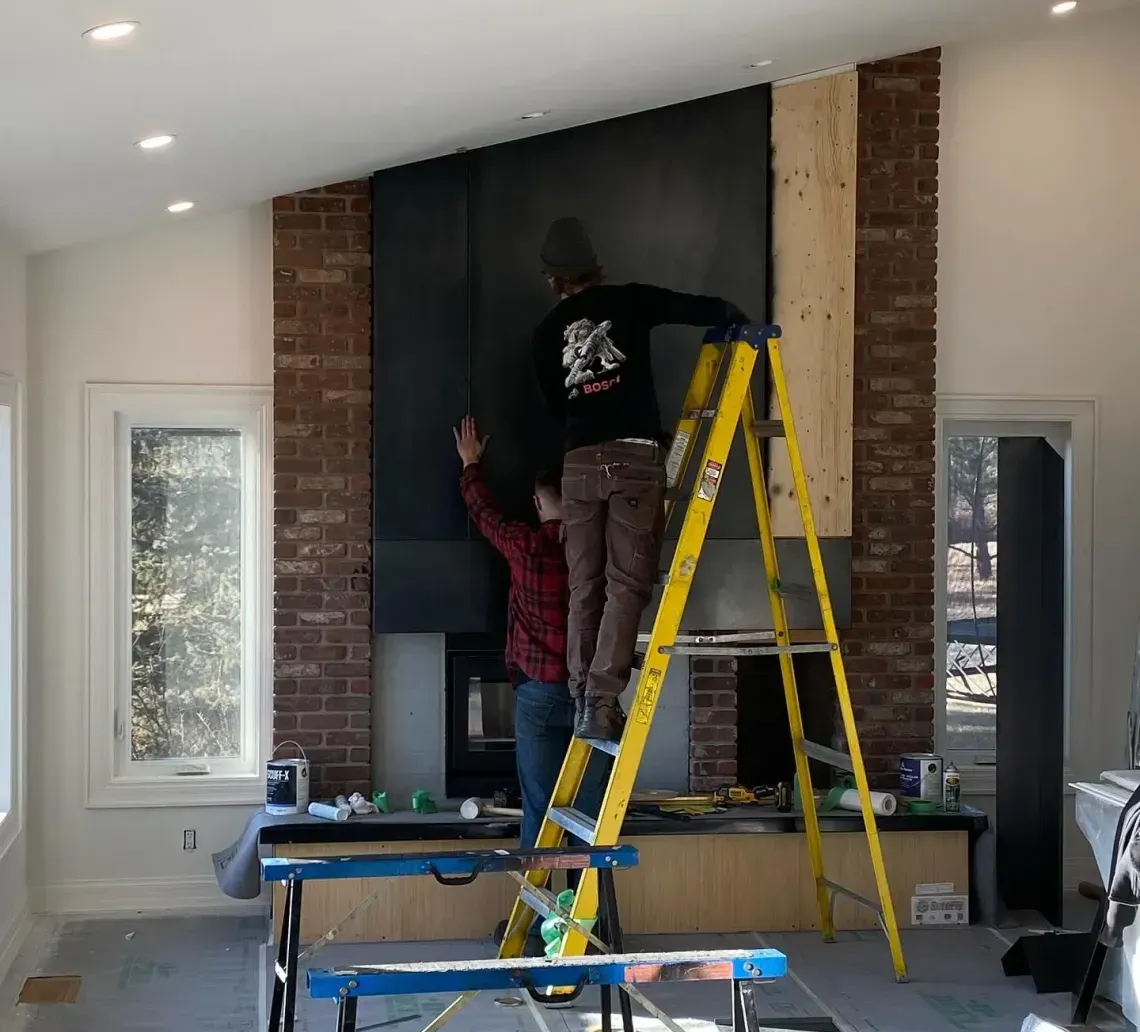 Two people work on a brick fireplace renovation, with one person on a yellow ladder installing large, dark panels.