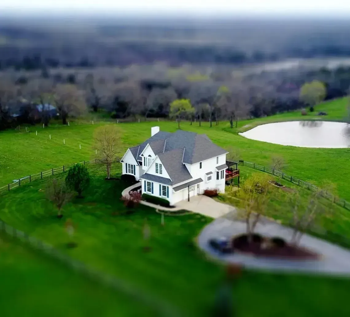 A white house sits in a grassy field next to a small circular pond, viewed from above with a tilt-shift focus effect.