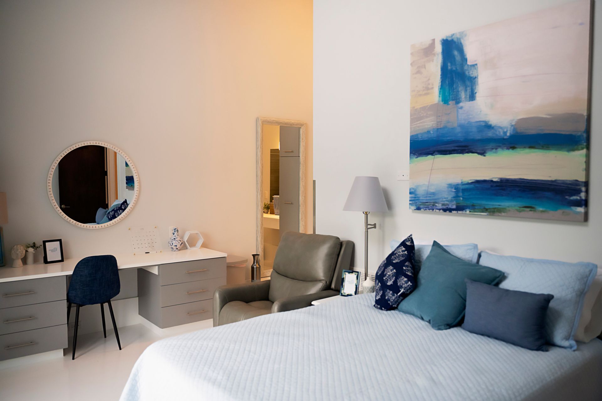 A bedroom with a bed , chair , desk , mirror and painting on the wall.