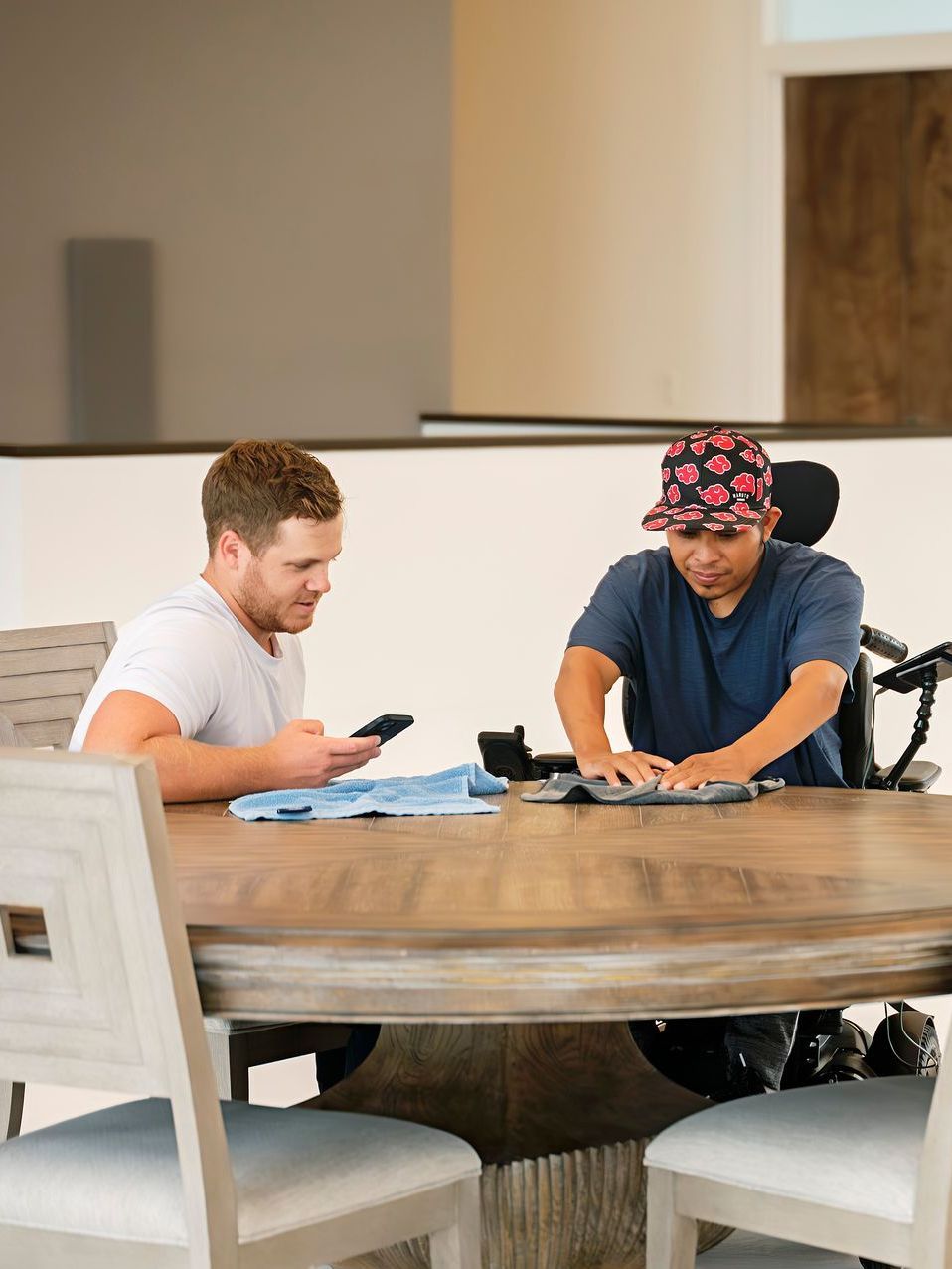 Two men are sitting at a table looking at their phones . one of the men is in a wheelchair.