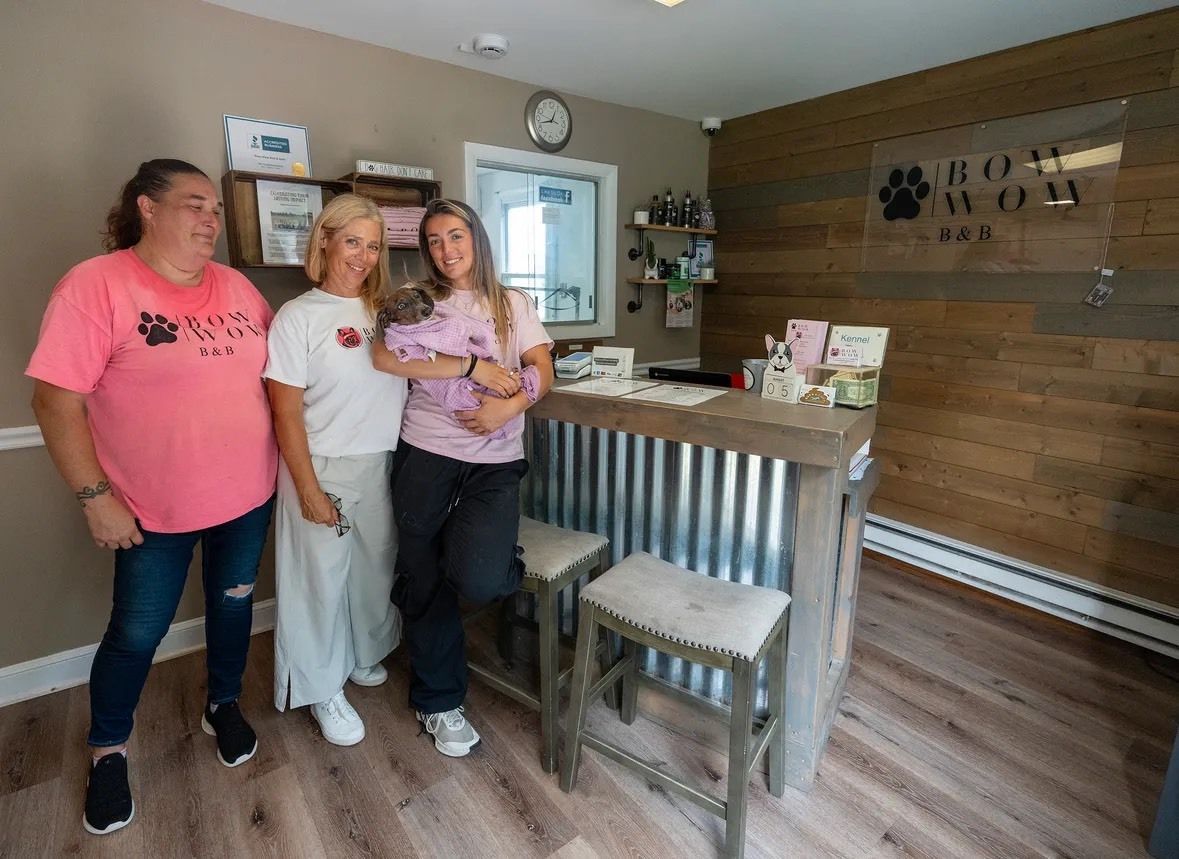 Three people stand behind a wood and metal reception desk in a pet groomer’s office. One person holds a small dog.