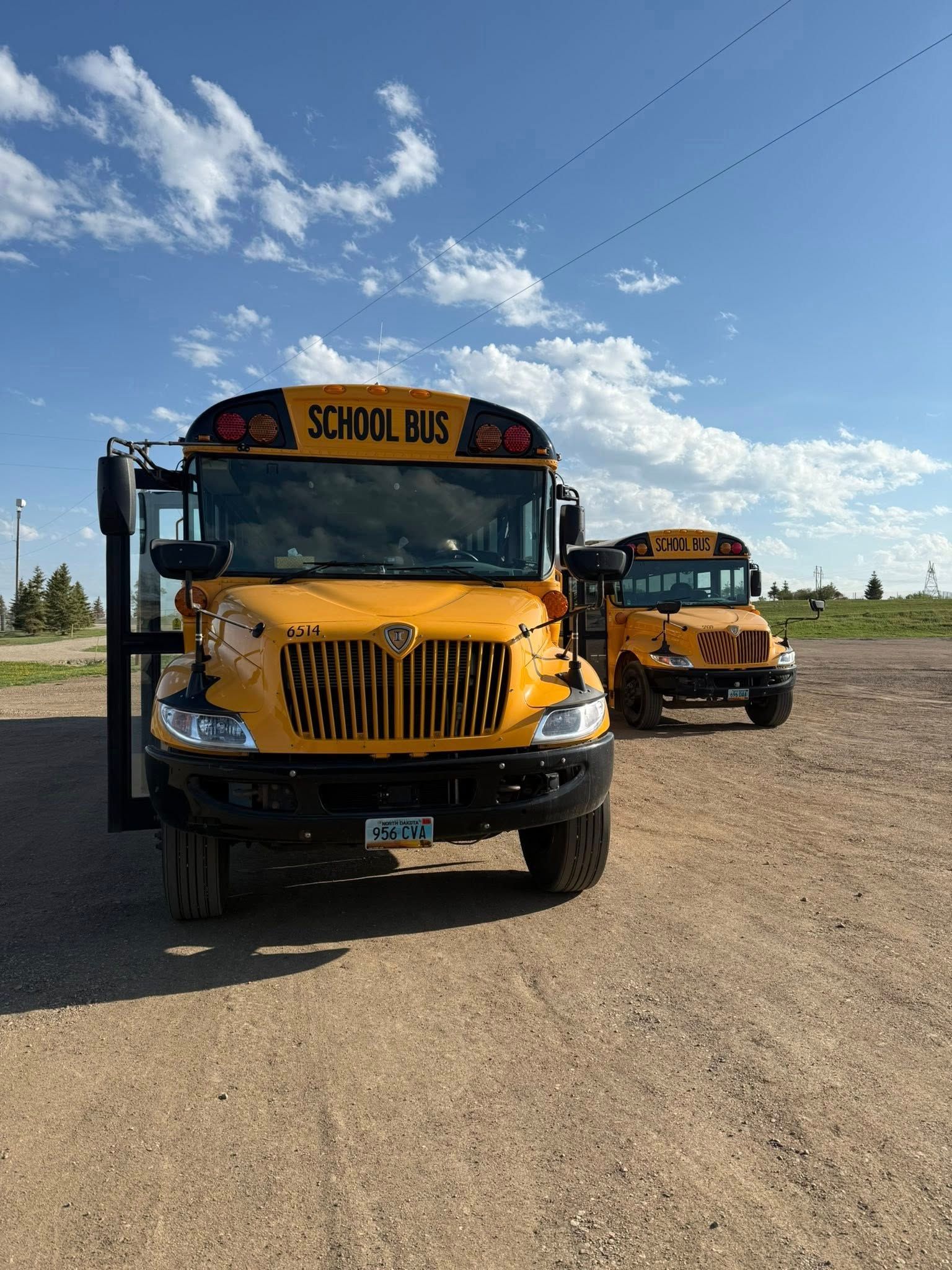 Two yellow school buses parked on a dirt lot under a blue sky with some clouds.