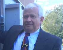 Man in a dark suit and colorful tie standing outdoors beside a blue structure, smiling slightly.