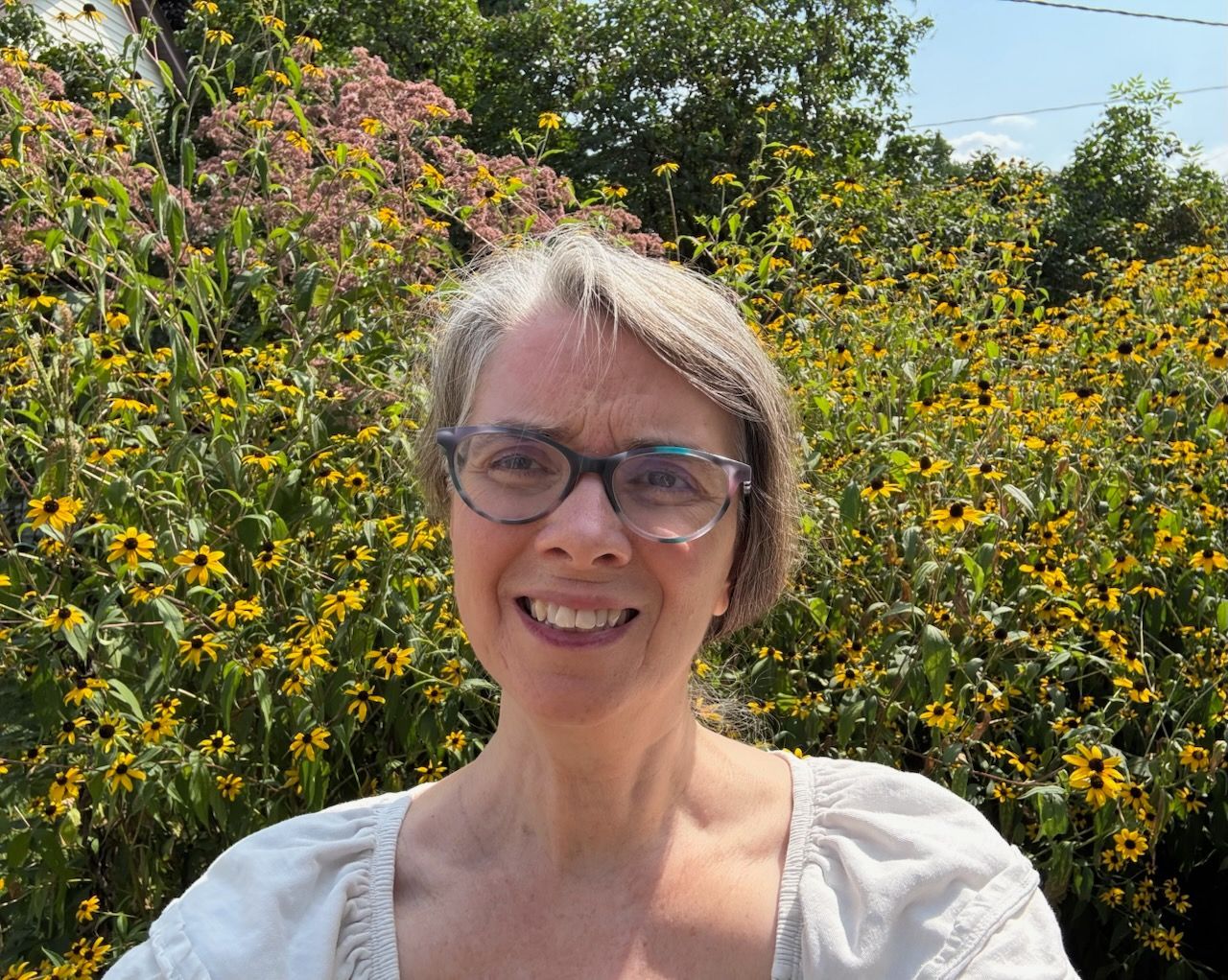 Smiling person in glasses standing outdoors in front of yellow wildflowers under bright sunlight