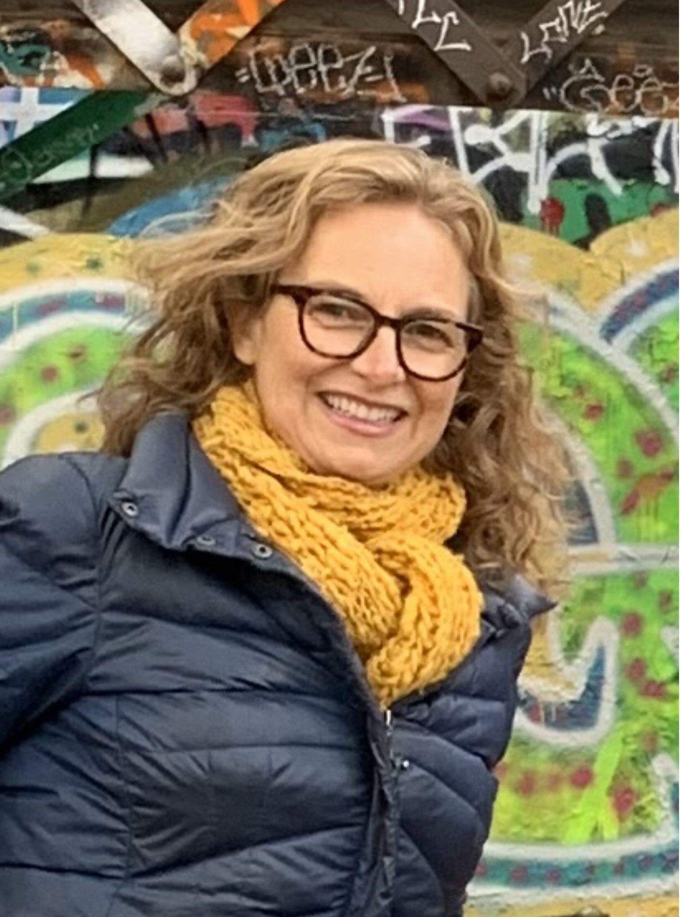 Smiling woman in glasses and a yellow scarf standing before colorful graffiti wall
