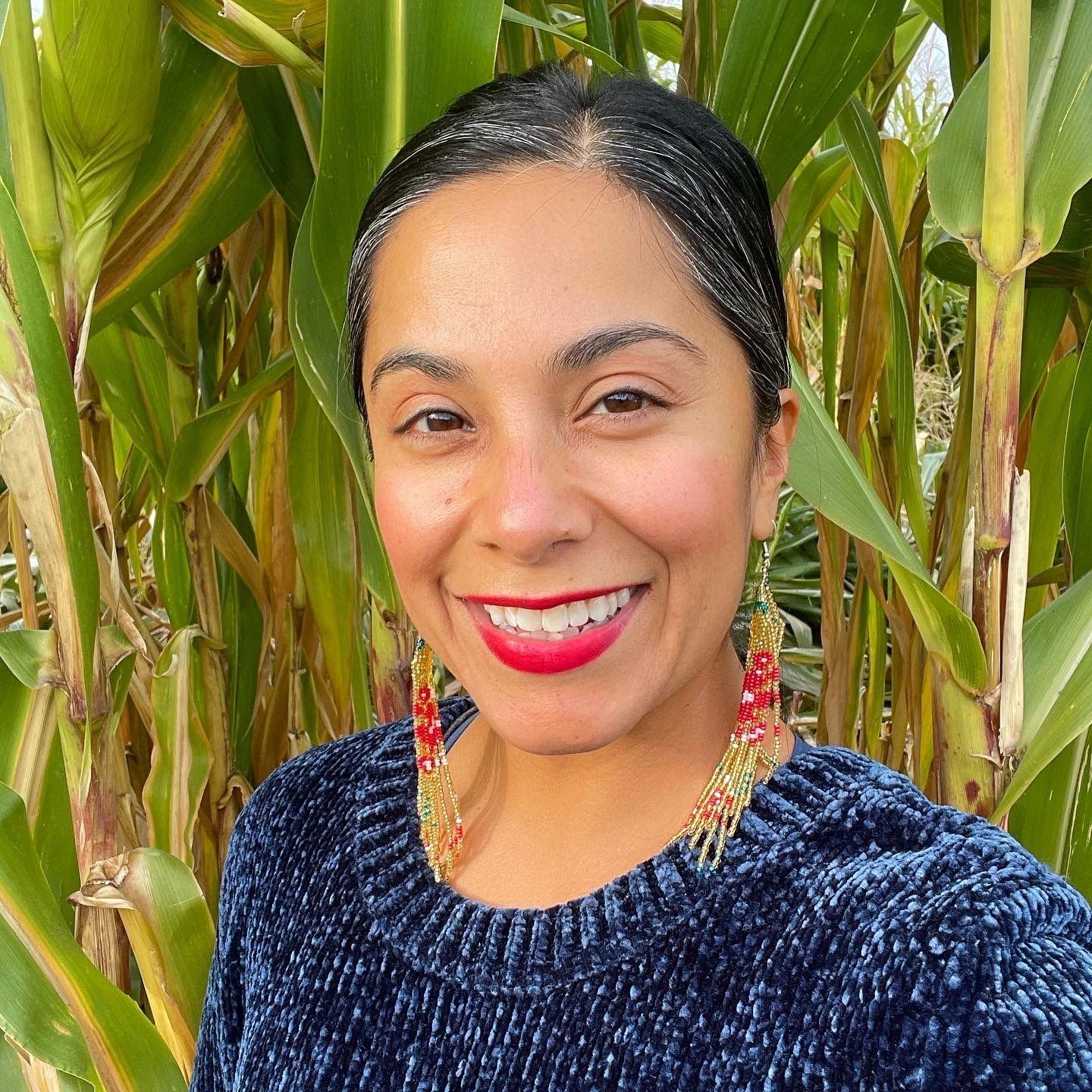 Smiling woman in a navy sweater standing in front of tall green corn plants.