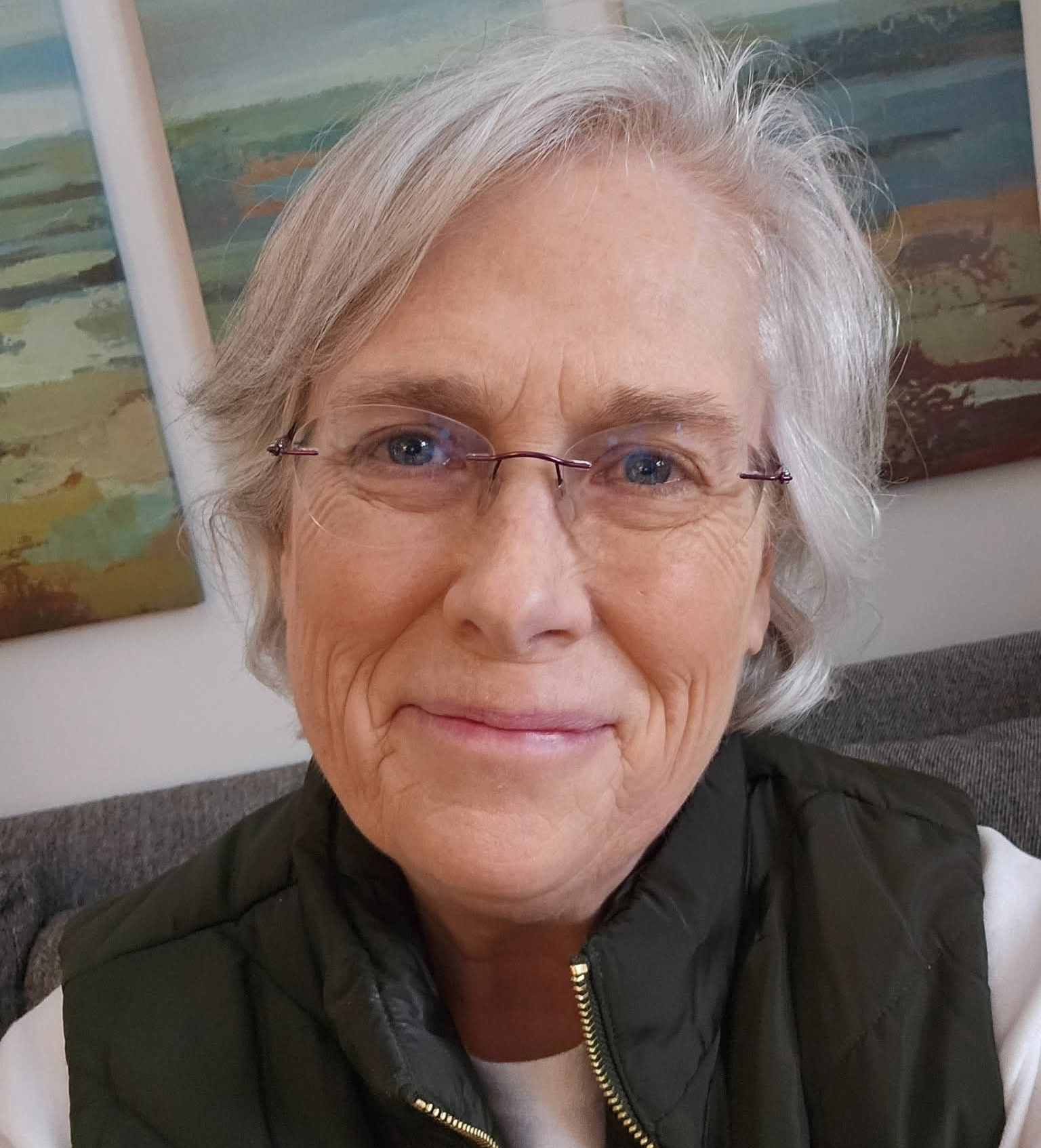 Smiling person with short white hair and glasses indoors, wearing a dark vest in front of framed wall art