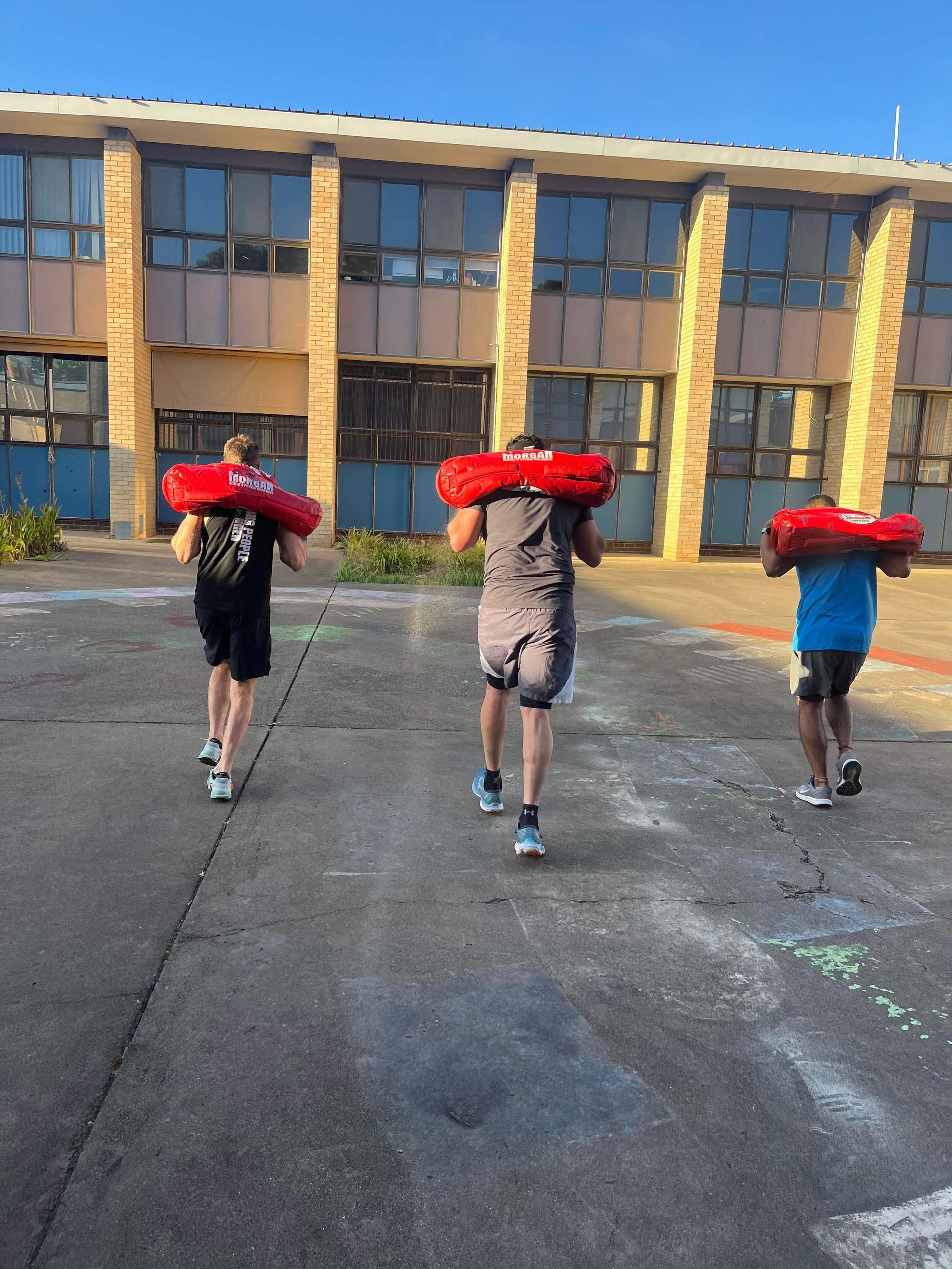 Three men are carrying red bags on their shoulders in front of a building.