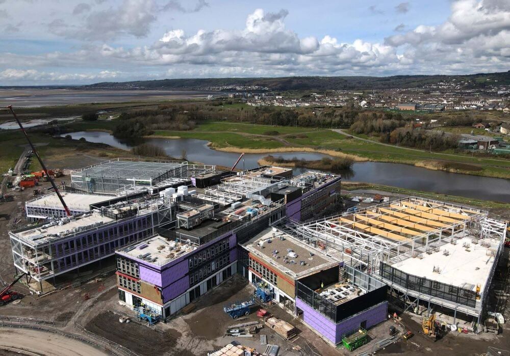 Aerial view of a multi-building construction site near a river and marshland, with purple accents and cranes.