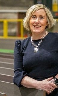 Woman with blonde hair wearing a black dress and silver necklace, smiling in a factory setting.