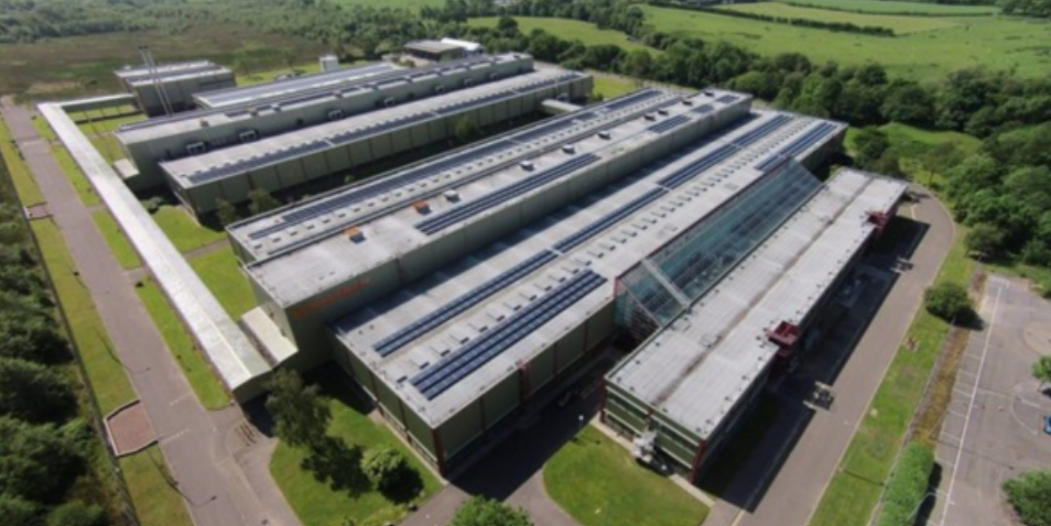 Aerial view of a large, modern industrial building with multiple flat roofs and solar panels, surrounded by greenery.