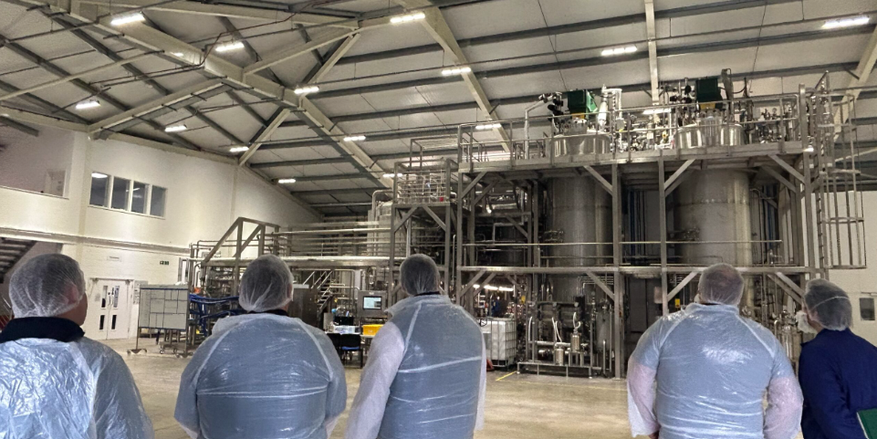 People in protective gear standing in front of stainless steel industrial tanks inside a large factory.