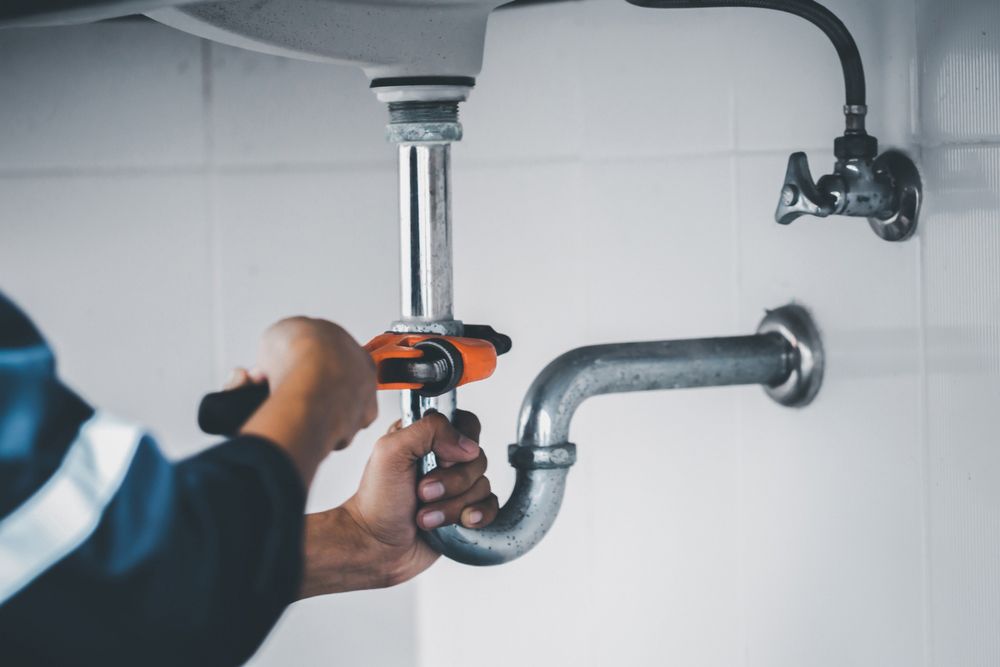 Plumber using a wrench to tighten a pipe under a sink with metal fittings and white tiled wall.