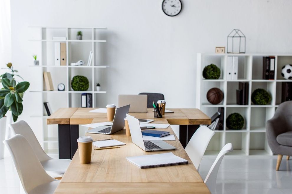 Ufficio luminoso e moderno con un lungo tavolo in legno, computer portatili e scaffali bianchi.