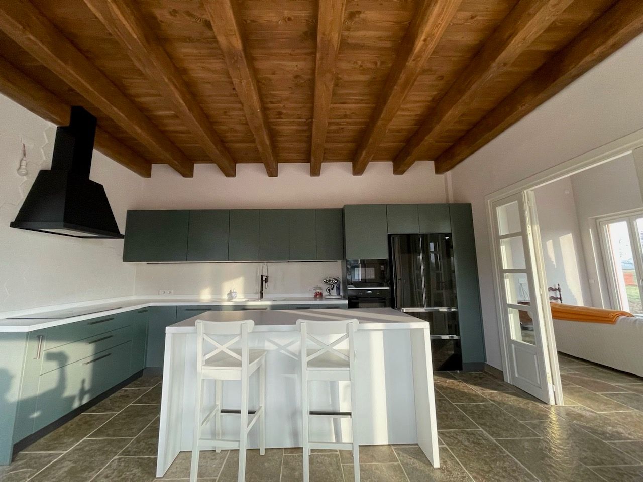 Cucina con mobili color verde acqua, soffitto con travi in legno, isola bianca con sgabelli ed elettrodomestici neri.