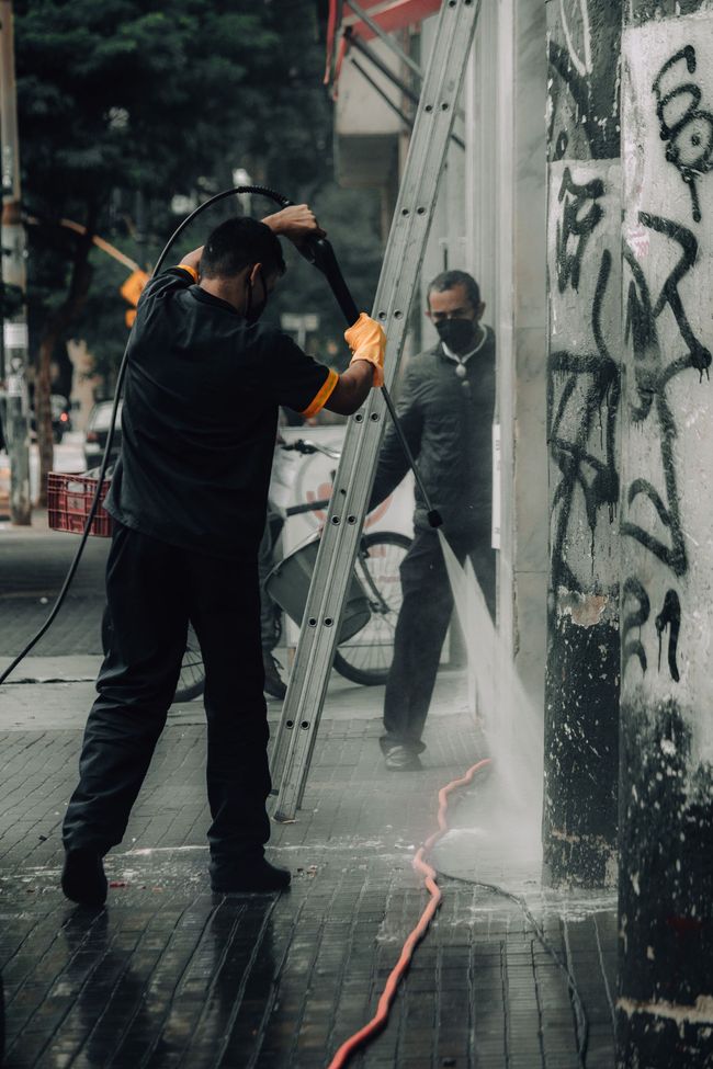 Worker power-washing a graffiti-covered wall on a rainy sidewalk, with water spraying and a ladder nearby