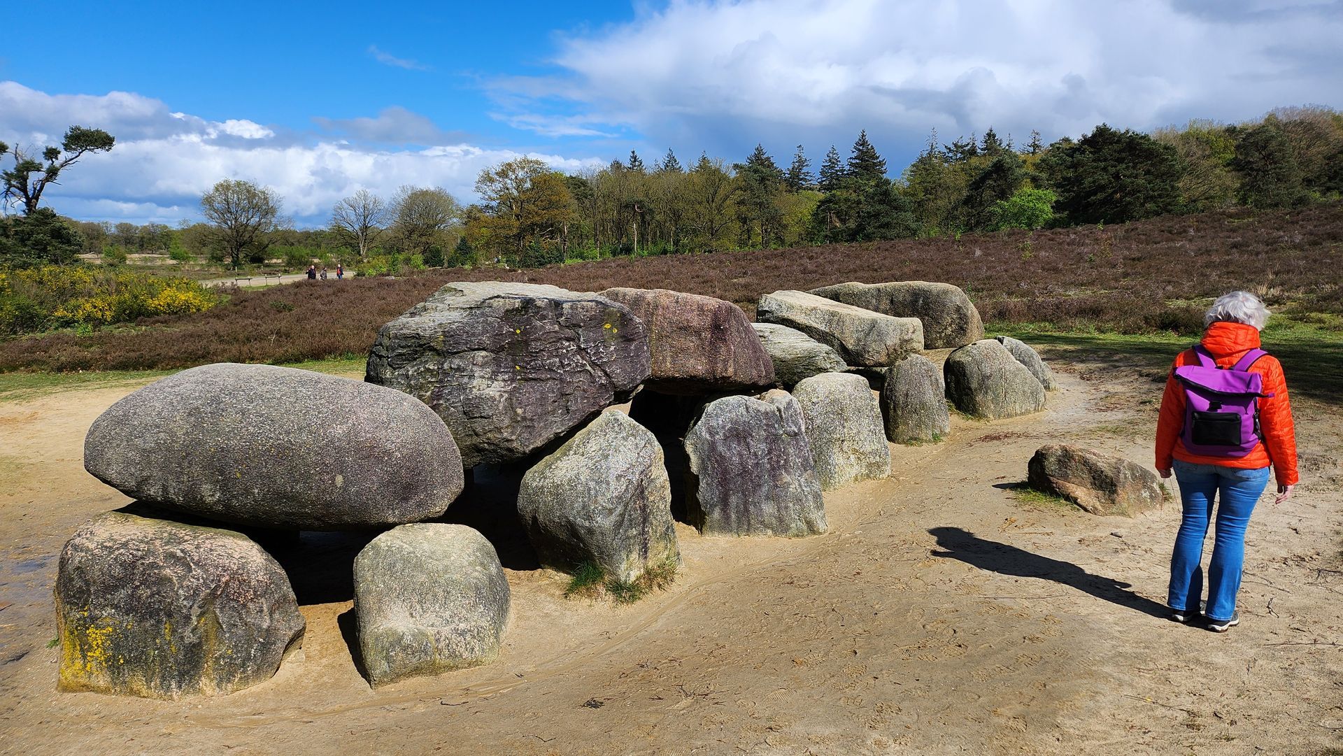 Een persoon met een oranje jas en een paarse rugzak bekijkt een stenen dolmen in een zonnig, met heide begroeid landschap.