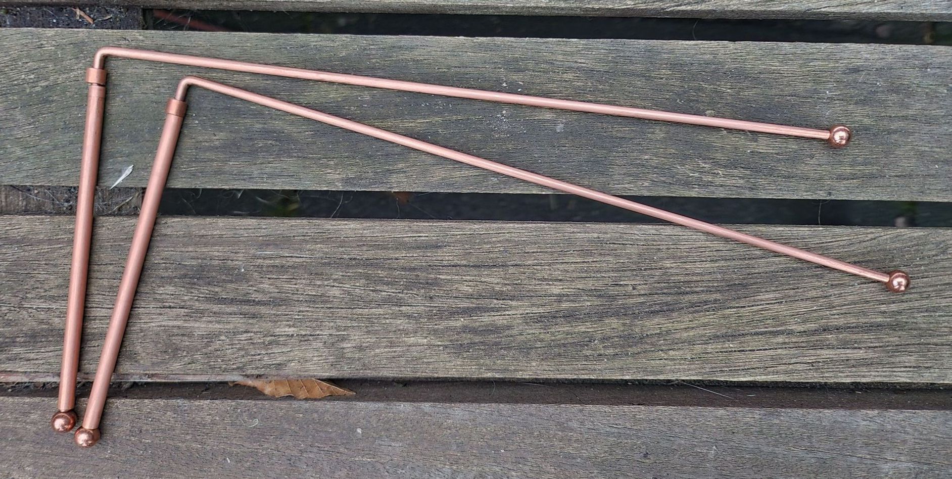 Twee L-vormige koperen wichelroeden met afgeronde uiteinden liggen op een verweerde houten ondergrond.