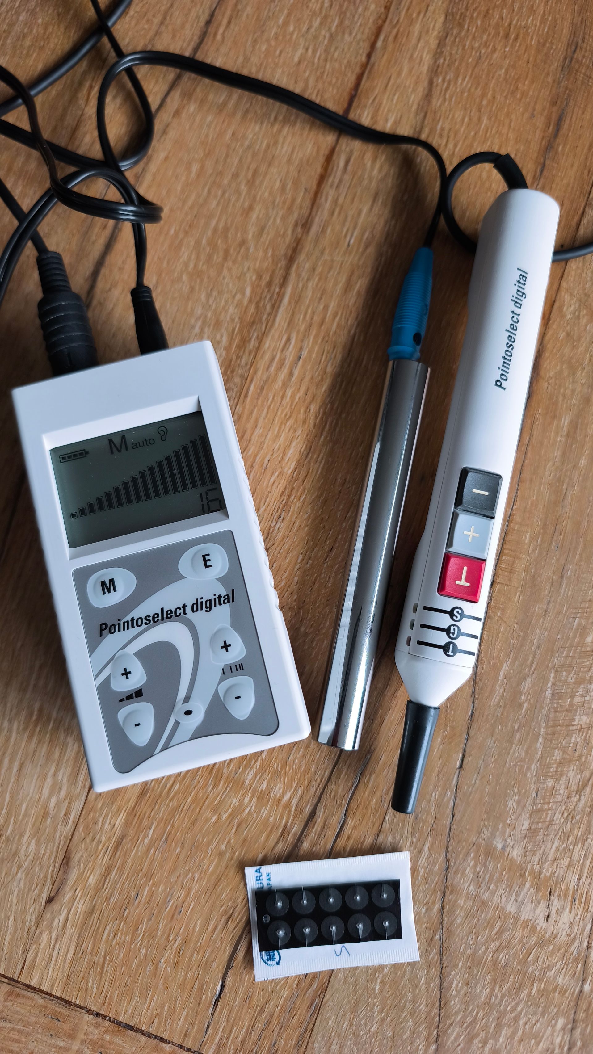 Een grijs elektro-acupunctuurapparaat met een digitaal scherm, een metalen sonde, een handstimulator en een set elektroden.
