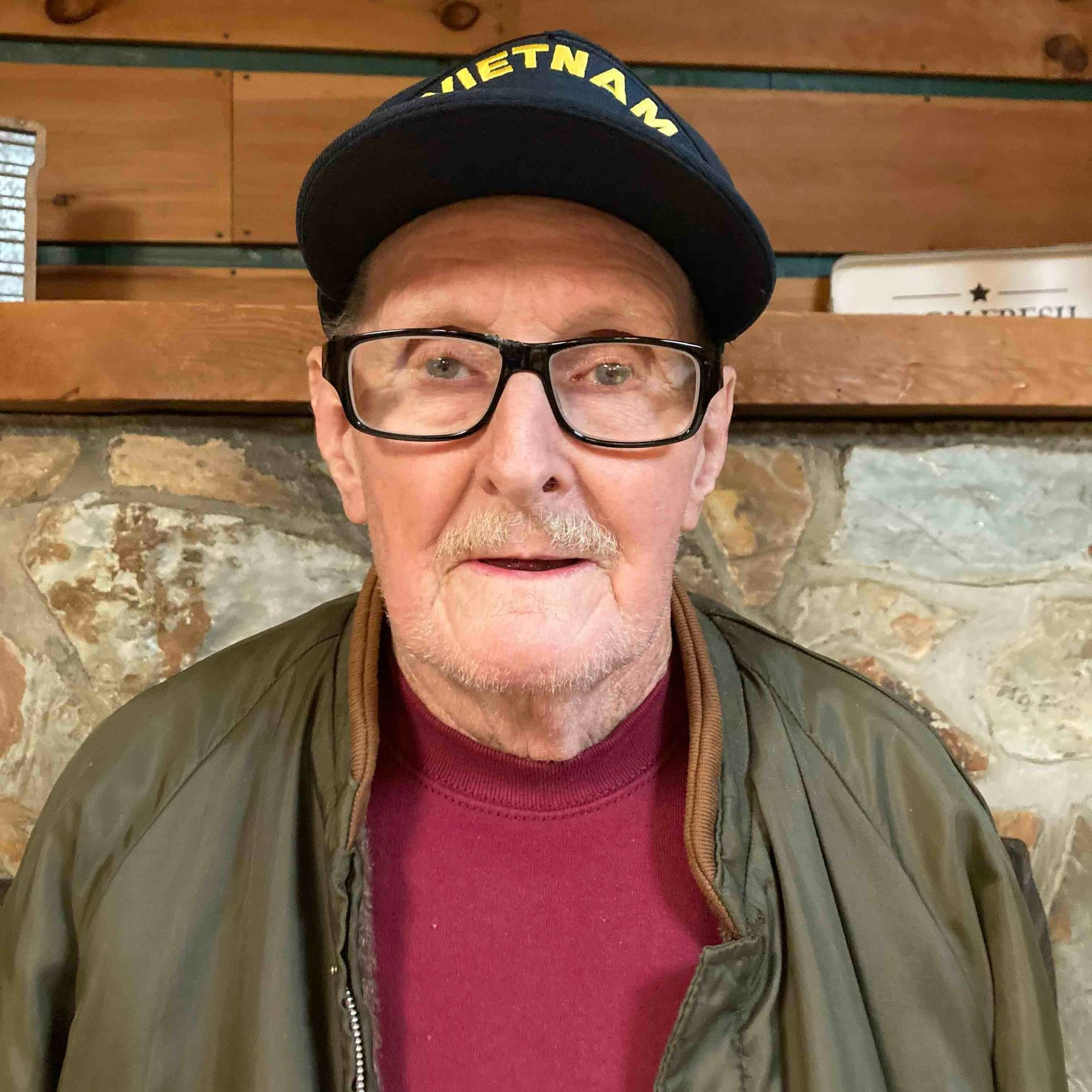 An older man wearing a hat and glasses is standing in front of a stone wall.