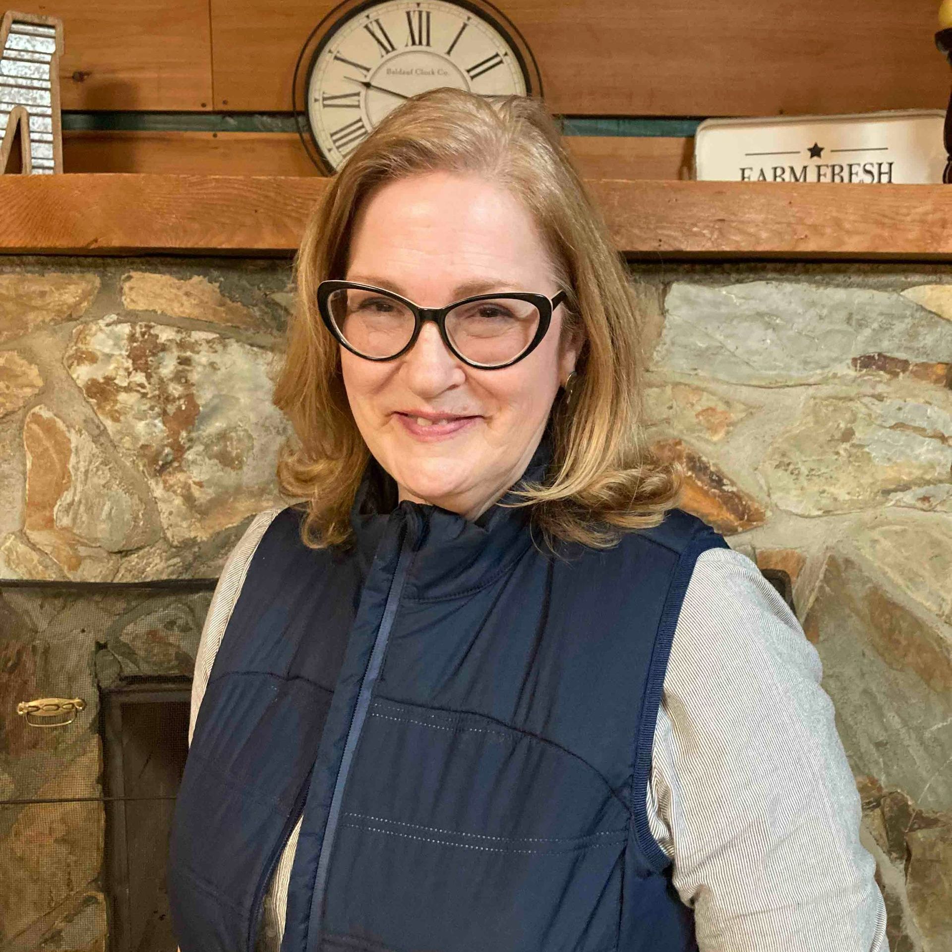 A woman wearing glasses and a vest is standing in front of a fireplace.