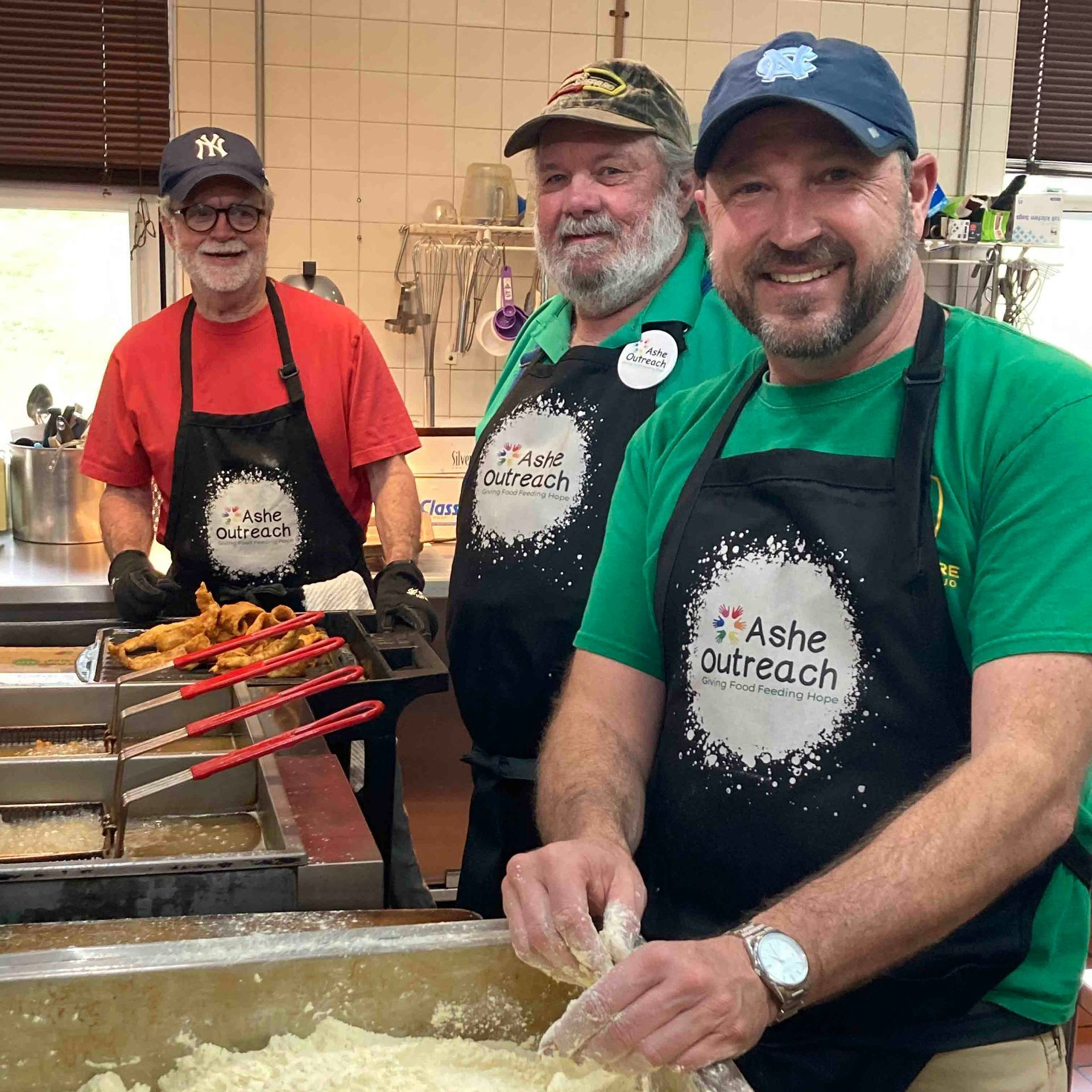 Three men are standing in a kitchen wearing aprons that say ash outreach